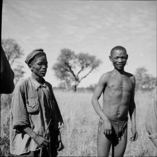 "Short /Qui" wearing Western clothes, standing, holding a bow, with another man standing next to him