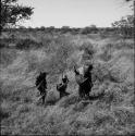 Three people carrying possessions on their backs, walking through the veld