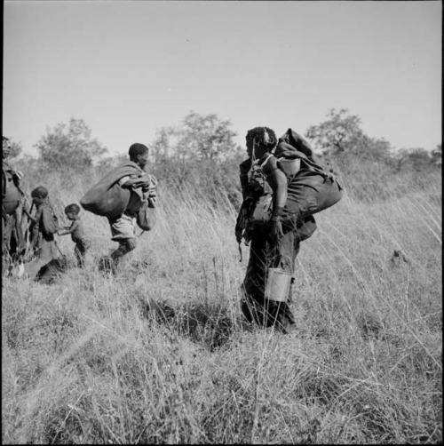 Woman standing, carrying possessions on her back, holding a tin bucket, with other people standing in the background