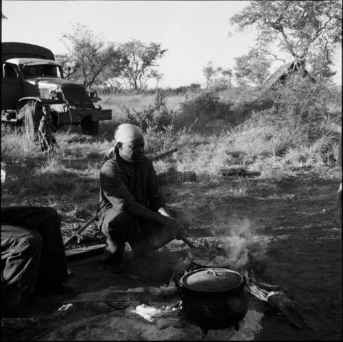 Man squatting, stirring food in a pot on a fire