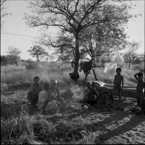 Group of people sitting and standing around a fire, with belongings hanging in a tree behind them