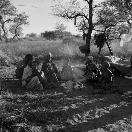 Group of people sitting around a fire, with belongings hanging in a tree behind them