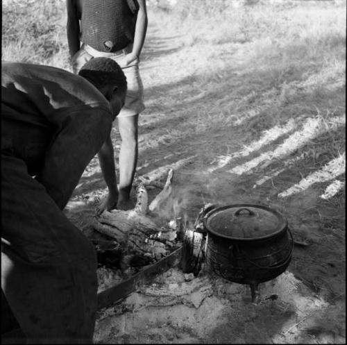 Expedition member leaning over, looking at a piece of burning wood, with an iron pot on the fire next to him