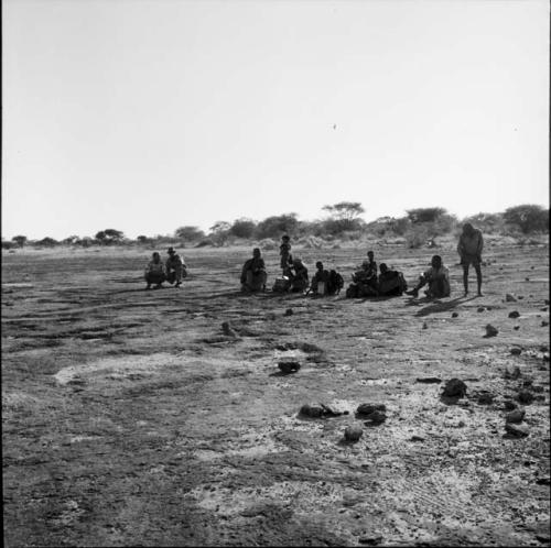 Group of people, including /Ti!kay, sitting, with a bucket and enamel cups on the ground next to them