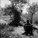 Man sitting, with a woman scooping something out of a bucket behind him, shelter skerm in the background