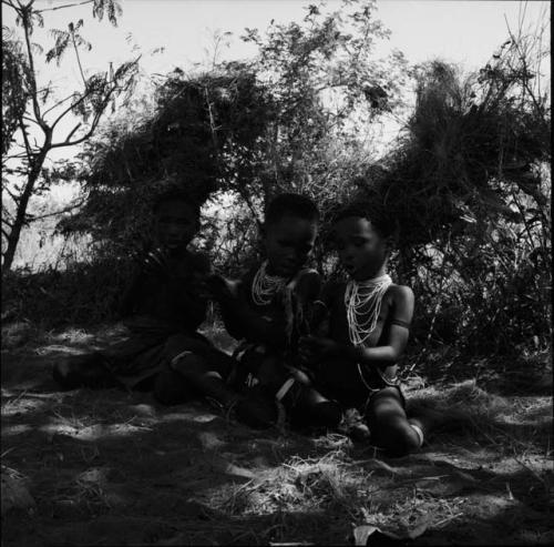 Three children sitting in the shade, clapping and singing, with two shelter skerms behind them