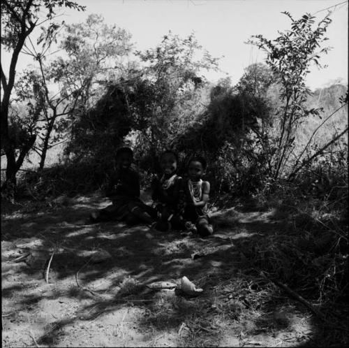 Three children sitting in the shade, clapping, one singing, with two shelter skerms behind them