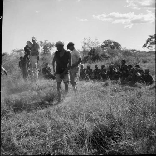 Two men dancing in a daytime dance, with a group of women sitting in a line, clapping and singing, in the background, expedition member filming