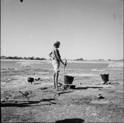 Man washing clothes at a pan, stirring one of two buckets on a fire, with an iron pot behind him, pan in the background