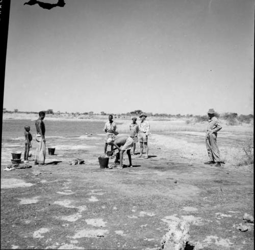 Man washing clothes at a pan, leaning over a bucket, with people standing near him, watching