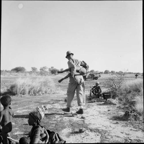 Nicholas England carrying a child across his back, with women and children sitting and standing near him