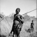 Man standing, holding an assegai, with other hunting equipment on his belt and over his shoulder, with a boy standing in the background