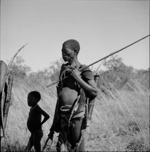 Man standing, holding an assegai, with other hunting equipment on his belt and over his shoulder, with a child standing behind him