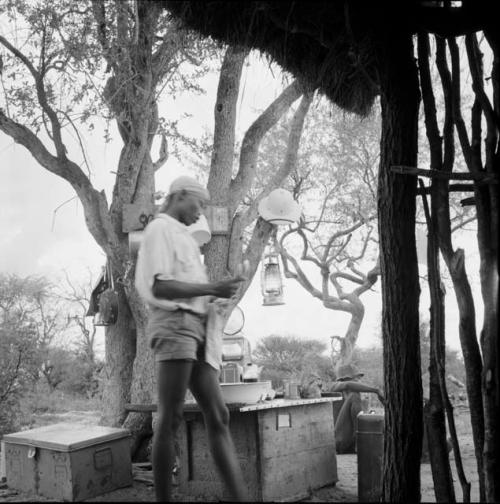 /Gaishay wearing Western clothes, standing outside the expedition hut, with a table and equipment on the ground next to him