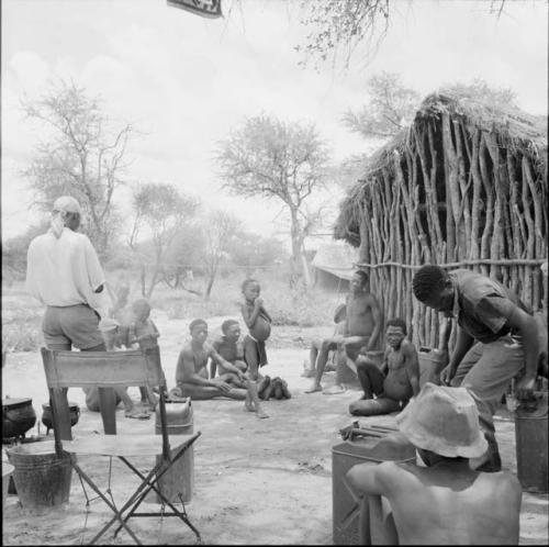 ≠Toma, /Ti!kay and /Gaishay with a group of people sitting and standing outside the expedition hut, with equipment on the ground near them