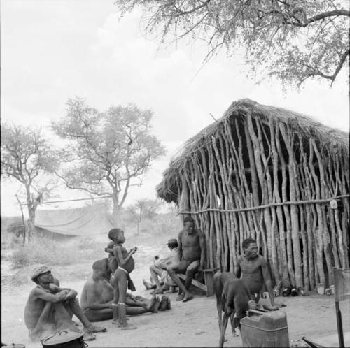 ≠Toma, /Ti!kay and /Gaishay with a group of people sitting and standing outside the expedition hut, with equipment on the ground near them