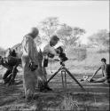 Man aiding a medicine man lying on the ground in trance, with John Marshall adjusting a camera, Laurence Marshall watching, people sitting and standing in the background