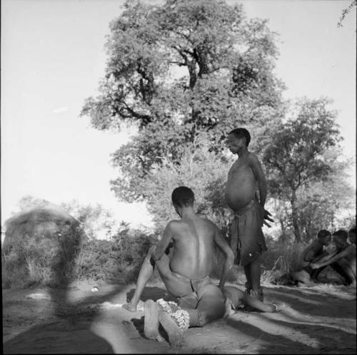 Man wearing dance rattles lying down in trance, with a man sitting next to him, another dancer standing behind them