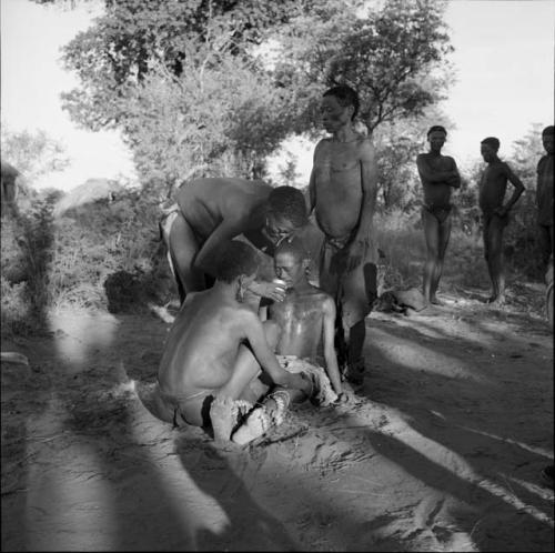 /Gunda in trance, sitting up, drinking from a cup held by another man, other men standing near them