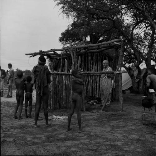 Group of children standing, looking at Laurence Marshall standing in the expedition hut, with expedition members standing, tent in the background