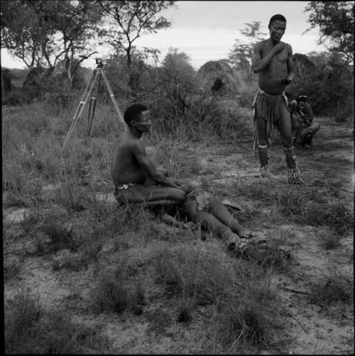 Man squatting, aiding a man lying on the ground in trance at the beginning of a daytime dance, with /Ti!kay wearing dance rattles, standing near them, a camera tripod and skerm in the background