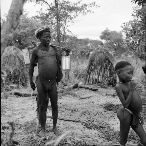 ≠Toma wearing a skin hat, carrying tin cans over his shoulder with carrying stick, with a boy walking next to him, partially constructed shelter skerms in the background