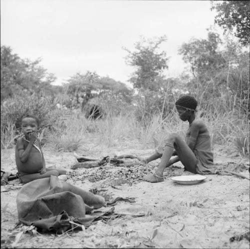 Woman sitting, cracking nuts, with a child sitting near her, chewing on a nut