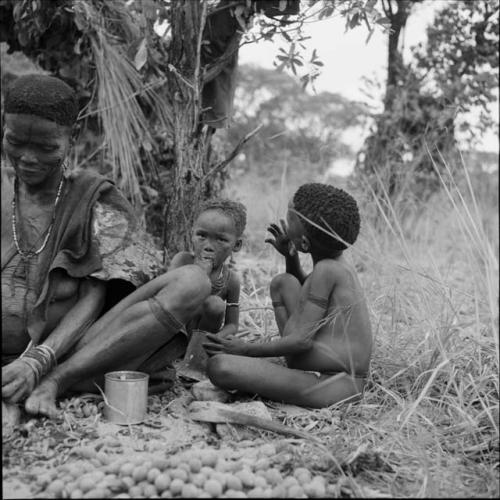 Woman sitting, cracking nuts, with two children sitting next to her, eating