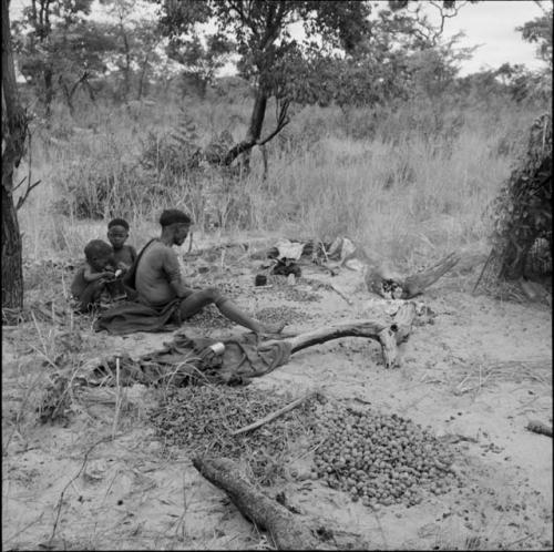 Woman sitting near a large pile of nuts, cracking nuts, with two children sitting next to her, eating