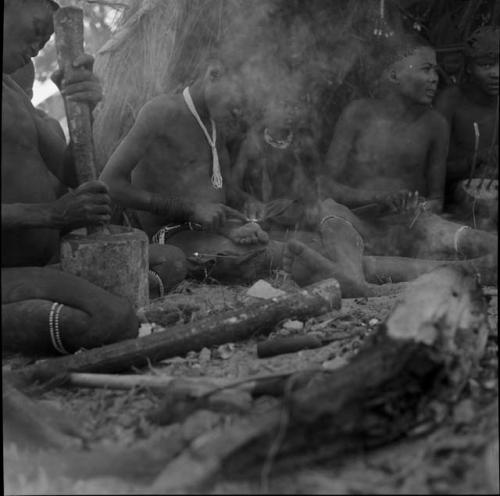 Five men sitting near a fire, one pounding with a mortar and pestle, two sharpening arrows, one playing a //guashi