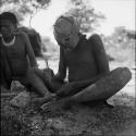 Man making an arrow point, with another man stirring a fire, sitting with him