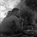 Man heating his awl in fire to make a hole for his adze, sitting with a group of men in front of a skerm