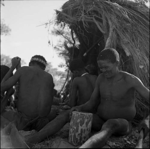 Dam holding a log, making a //guashi, sitting with a group of people in front of a skerm