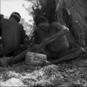 Dam working on a log with his adze, making a //guashi, sitting with a group of people in front of a skerm