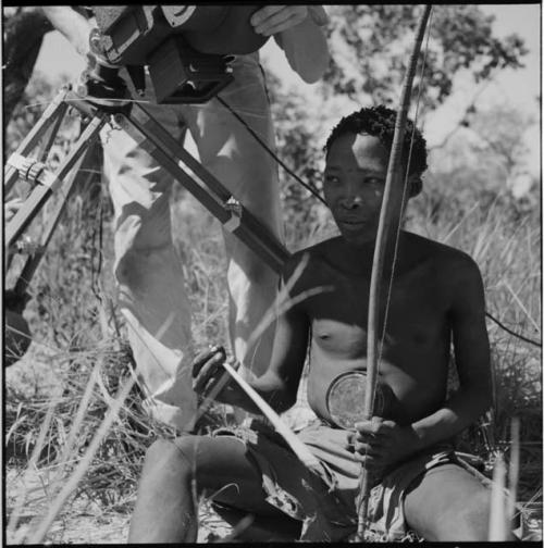 Man sitting, holding a musical bow, with an expedition member filming him