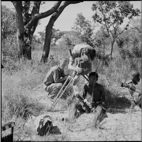 Man playing a musical bow, with an expedition member filming him, John Marshall squatting behind them