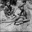 Two men sitting, one preparing to play a musical bow