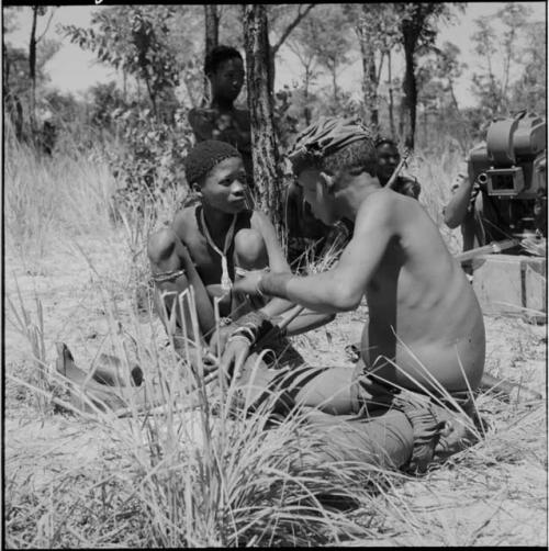 Two men sitting, one preparing to play a musical bow