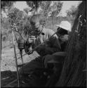 Nicholas England squatting near a microphone, examining a divided bow, with two expedition members sitting next to him