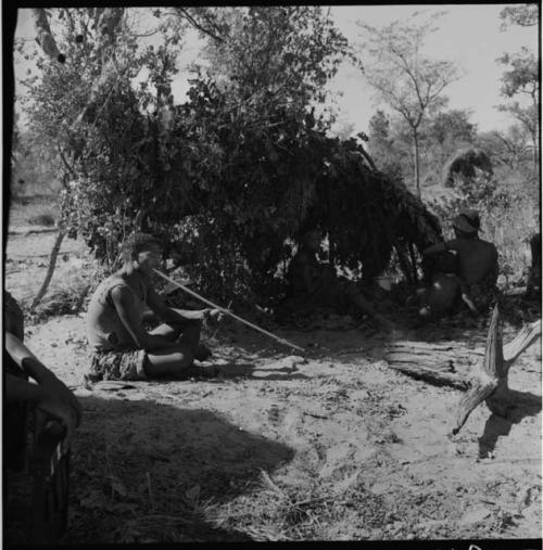 Man playing a musical bow, with a woman sitting inside a shelter skerm with her baby near him