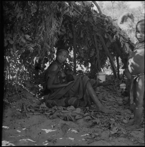 Woman sitting in a shelter skerm, holding a baby, with another child standing near her, enamel pot and bowl in the background