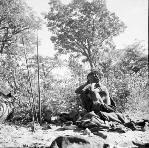 Woman wearing a kaross, sitting next to a blanket on the ground, three assegais and a bow stuck into the ground near her