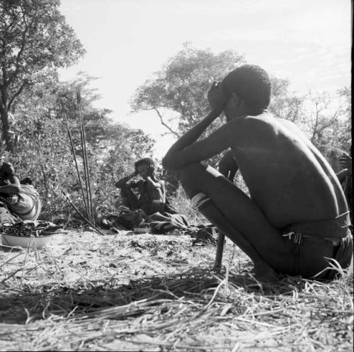 Man squatting, with a woman wearing a kaross, sitting next to a blanket on the ground in the background, three assegais and a bow stuck into the ground near her