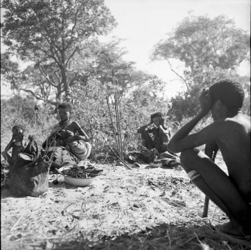 Man squatting, with two women and two children sitting in the background, a large bag and an enamel basin full of sinew on the ground near them