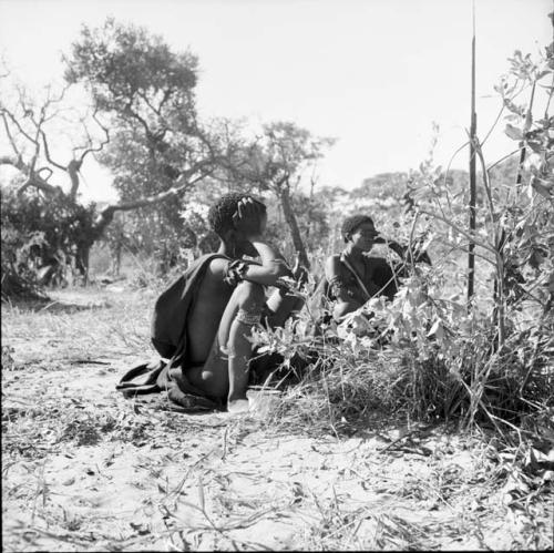 Woman wearing a kaross sitting, with a man sitting behind her, an assegai stuck into the ground near them