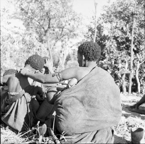 Two women with babies on their backs sitting, one reaching out to touch the other, view from behind