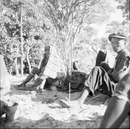 Woman wrapped in a striped blanket, with a child between her legs, sitting with a group of people, with assegais stuck into the ground near them