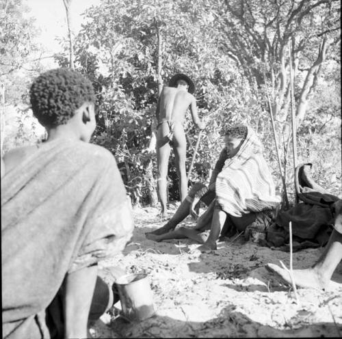 Woman wearing a kaross, sitting, with a woman wrapped in a blanket sitting with a child between her legs in the background, a man digging behind her