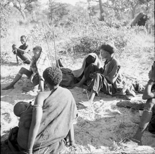 Group of people sitting, including a woman with a baby on her back in her kaross and a woman wrapped in a striped blanket with a child between her legs