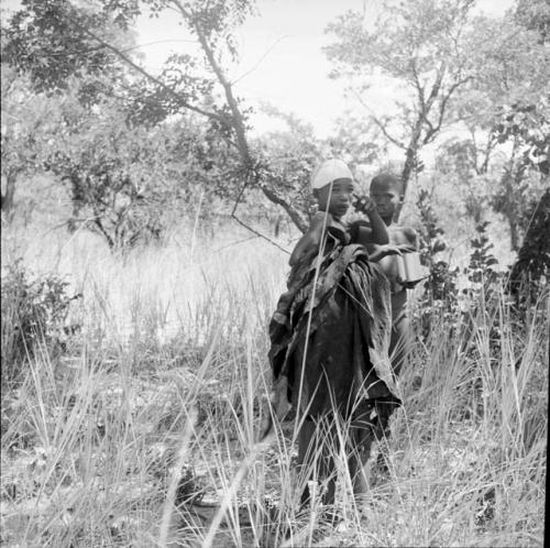 N!ai holding a large kaross, standing with a boy in the veld